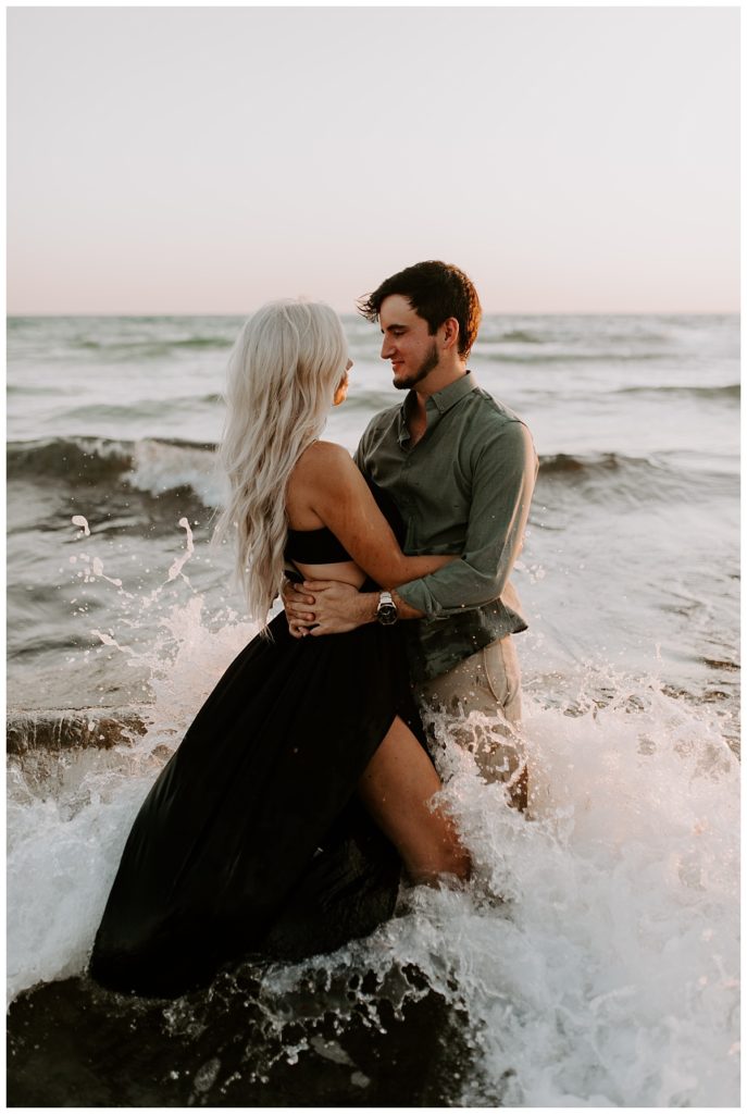 Woman in black dress and man in green shirt hold each other in waist deep water as a wave crashes around them - Rosemary Beach Photographer