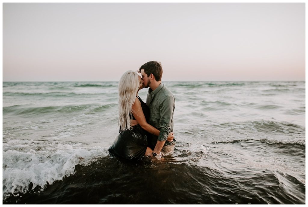 Man in green shirt and woman in black dress kiss each other while standing in the waist deep water on the beach - Rosemary Beach Photographer