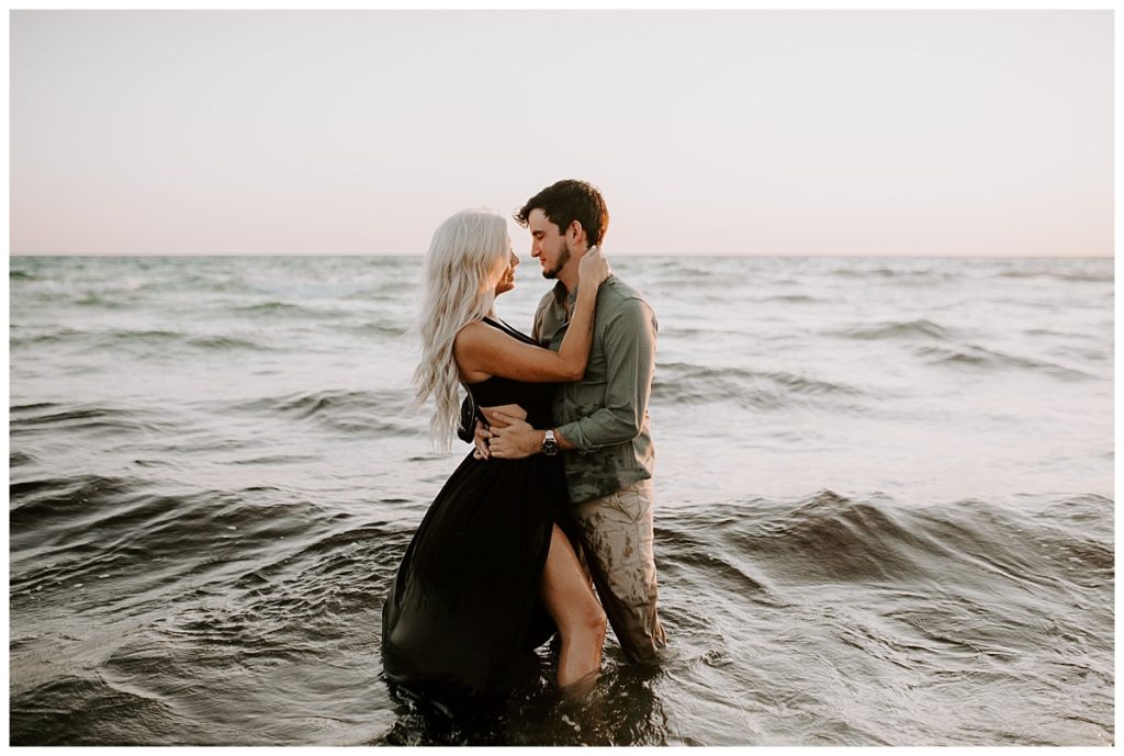 Woman in black dress and man in green shirt stand in waist deep water gazing into each others eyes