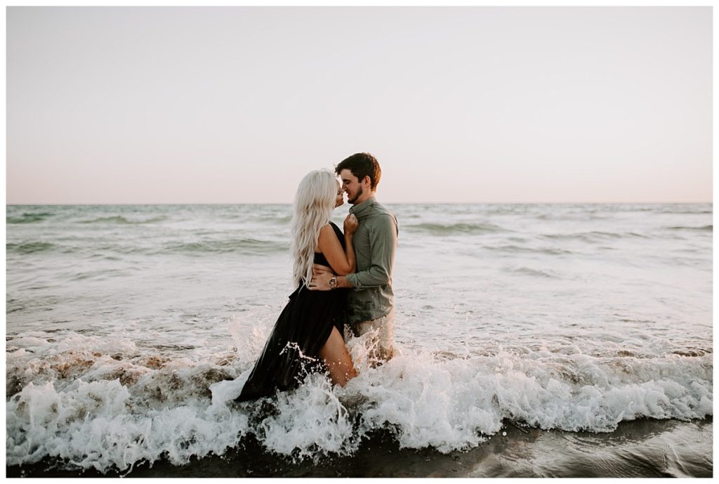 Woman in black dress and man in green shirt kiss in waist deep water while waves crash around them