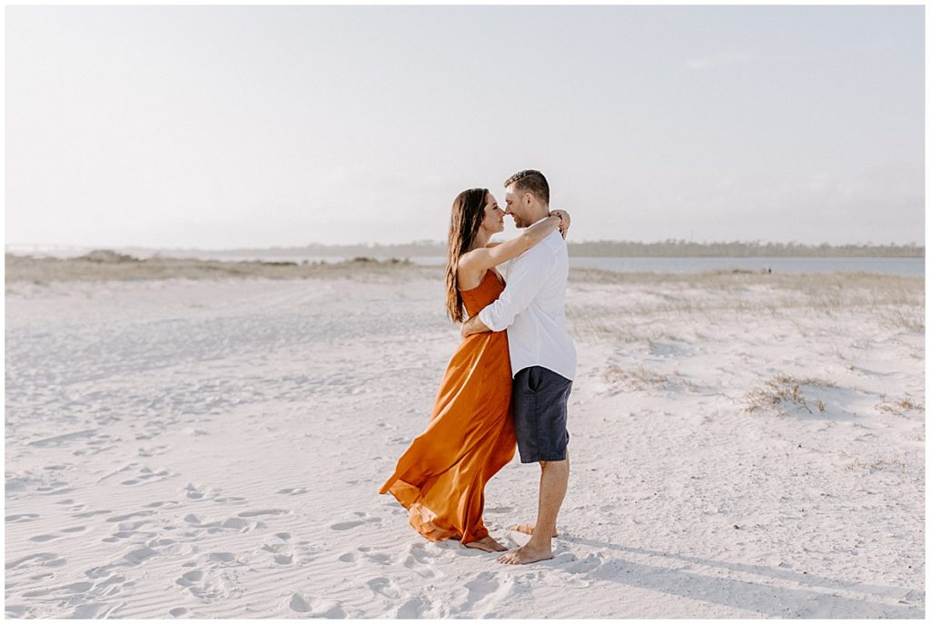 Couple - woman is dressed in rusty orange spagetti strap dressand man is in navy shorts with a white button down. They're holding each other on a white sand beach with a lake behind them.