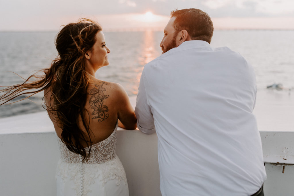 couple in wedding clothes looking at each other while the sun sets in front of them. They're on a boat.