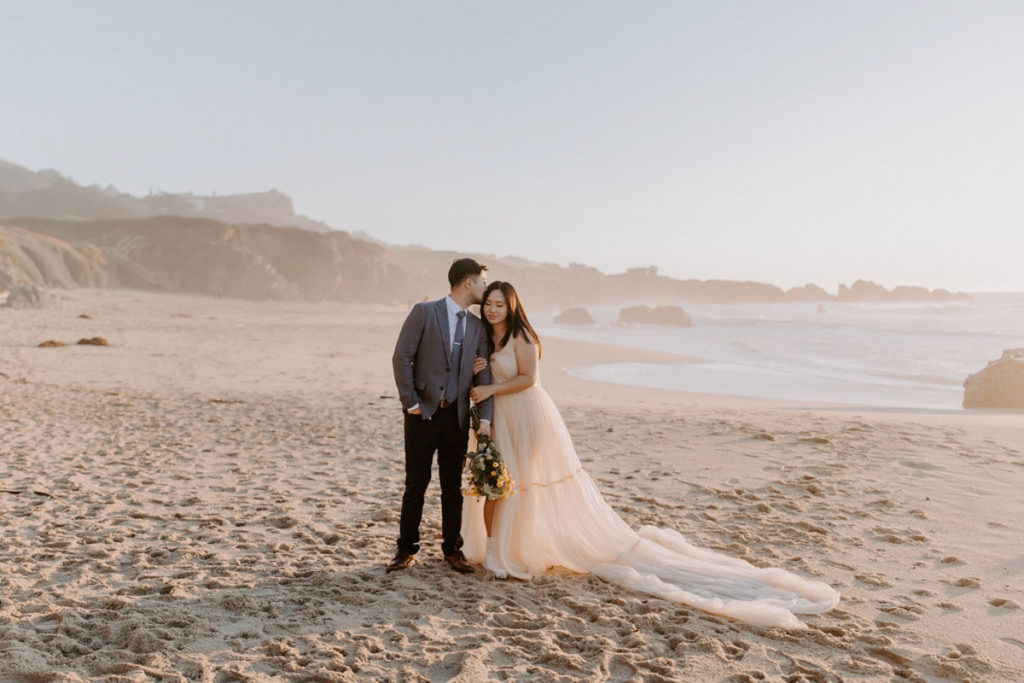 A couple is standing on a beach in their elopement attire. They are facing the camera, and the bride is hugging the groom's arm while he kisses her on the head.