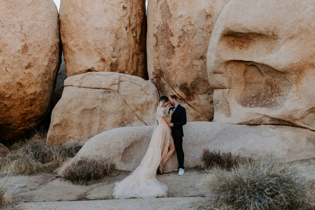 A couple is standing in front of boulders at Joshua Tree National Park, wearing elopement attire. They are facing each other, their foreheads together.