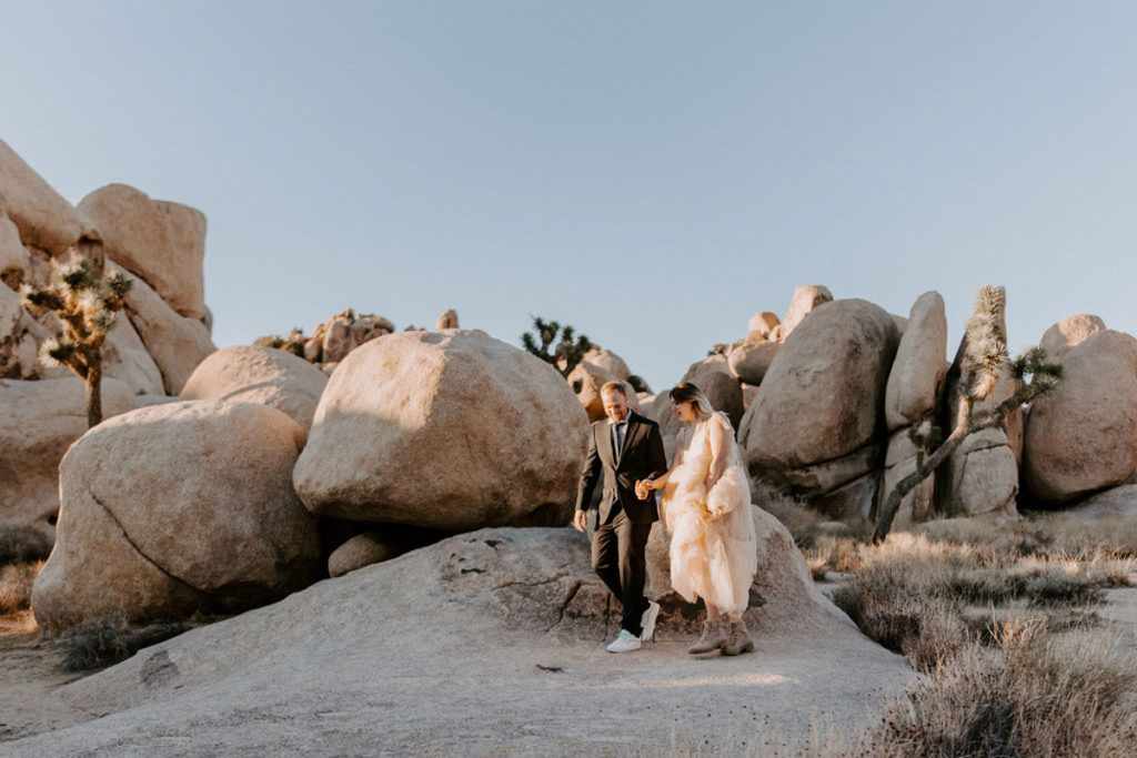 A couple is waling in front of boulders at Joshua Tree National Park. They are wearing elopement attire and holding hands, the groom waling ahead and leading the bride.