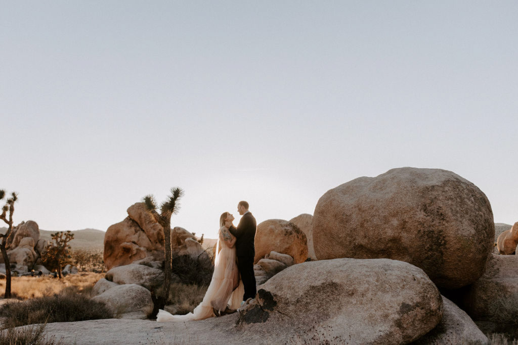 A couple is standing in front of boulders at Joshua Tree National Park, wearing elopement attire. They are facing each other, and the groom has his hands on the bride's face.