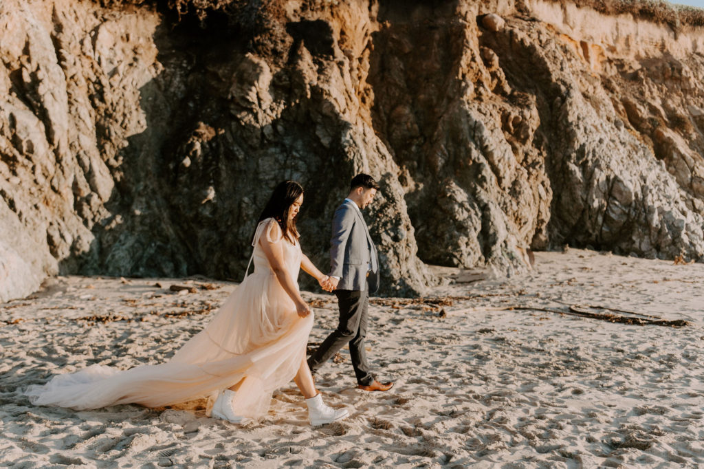 After traveling for their elopement in Big Sur, this couple is walking down the beach, wearing a wedding dress and a suit.